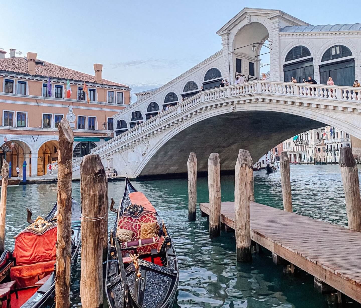 A low-angle shot of the white stone Rialto Bridge over the turquoise water of the Grand Canal. In the foreground, traditional black gondolas with ornate red and gold seating are moored to wooden pilings next to a small wooden pier.