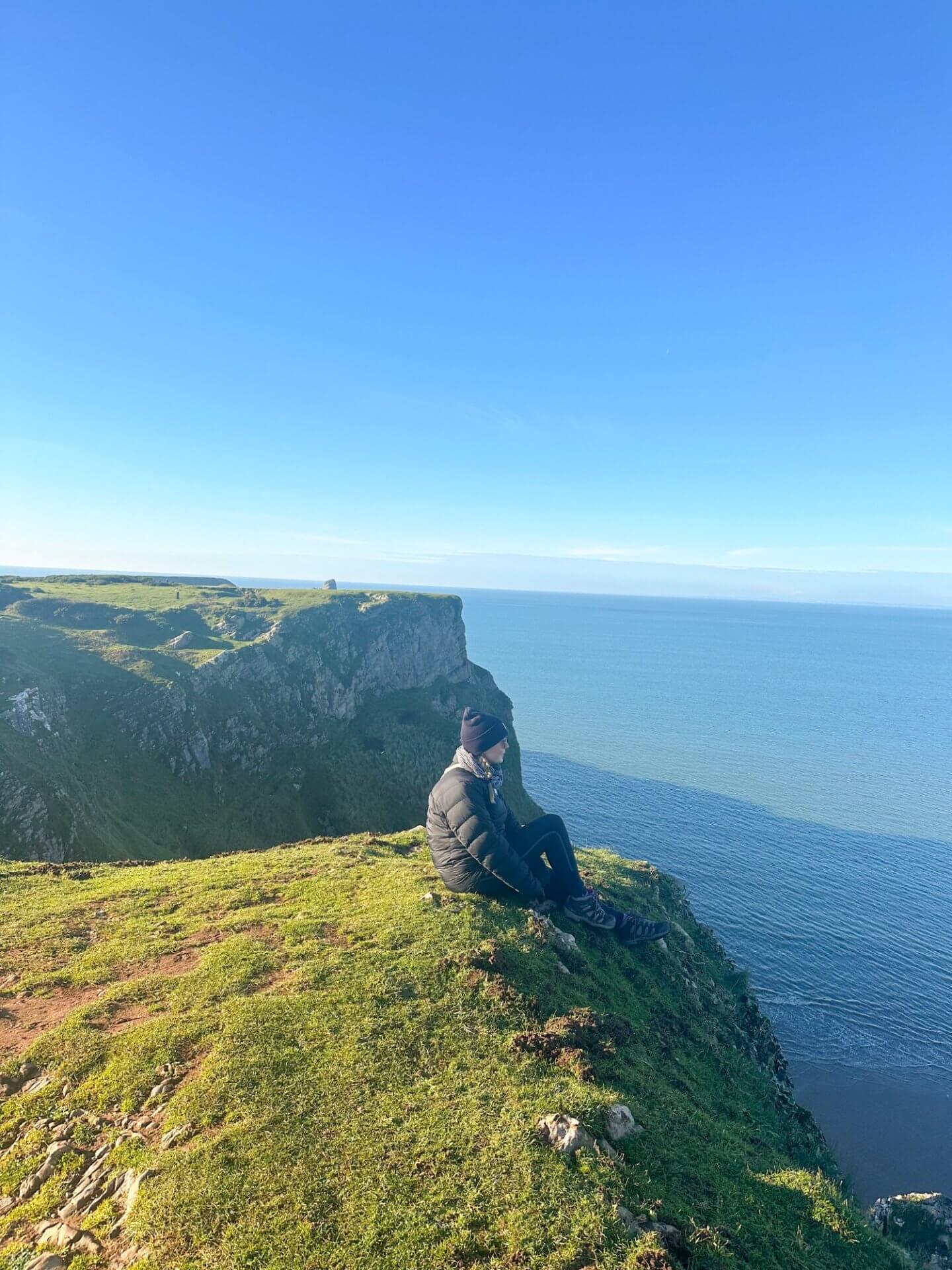 Image of a person sitting on a cliff looking over Rhossilli Bay, Wales, which is one of the best things to do in Gower