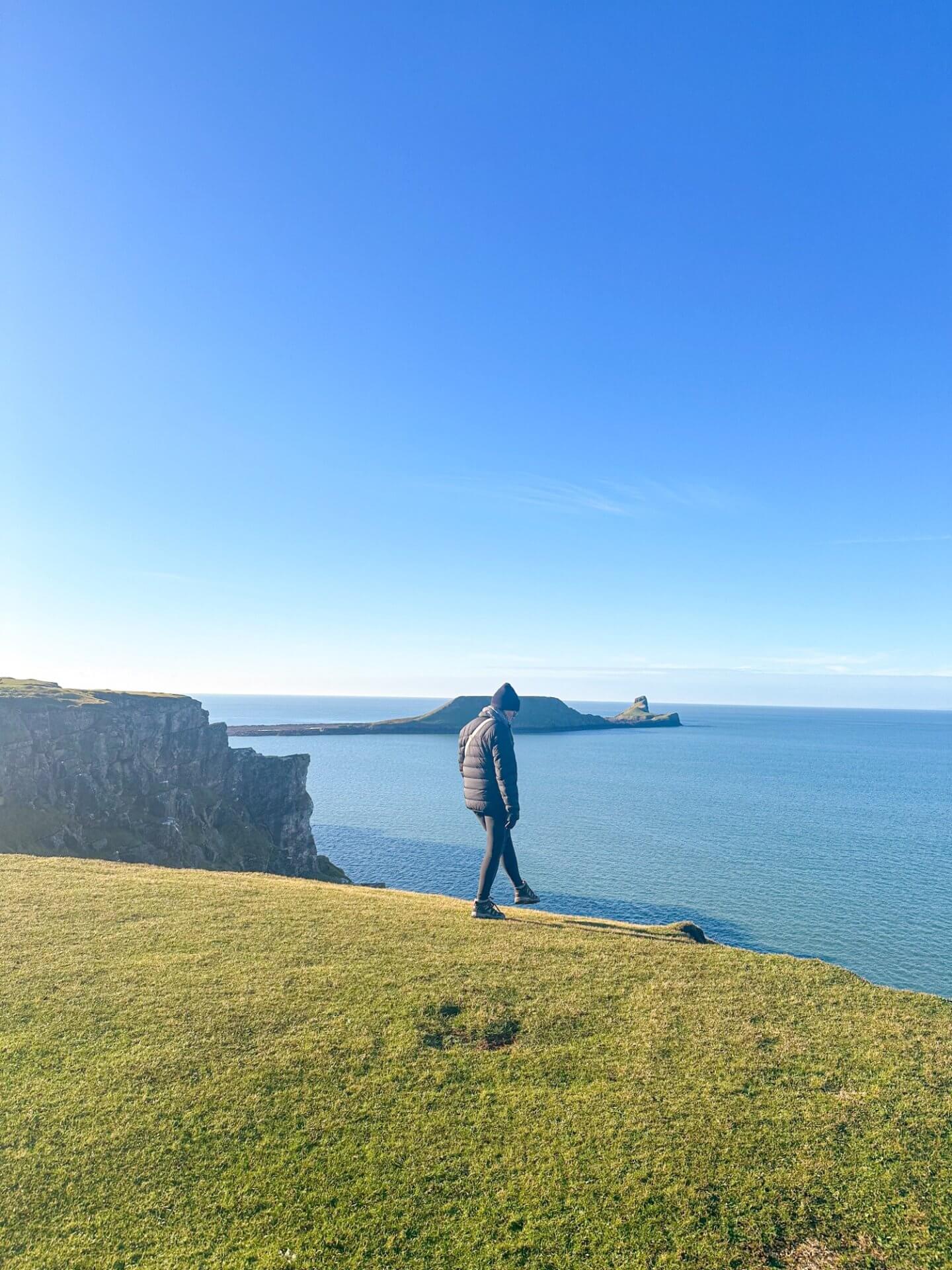 Image of a standing on the edge of a  cliff infront of Rhossilli Bay, Wales, which is one of the best things to do in Gower