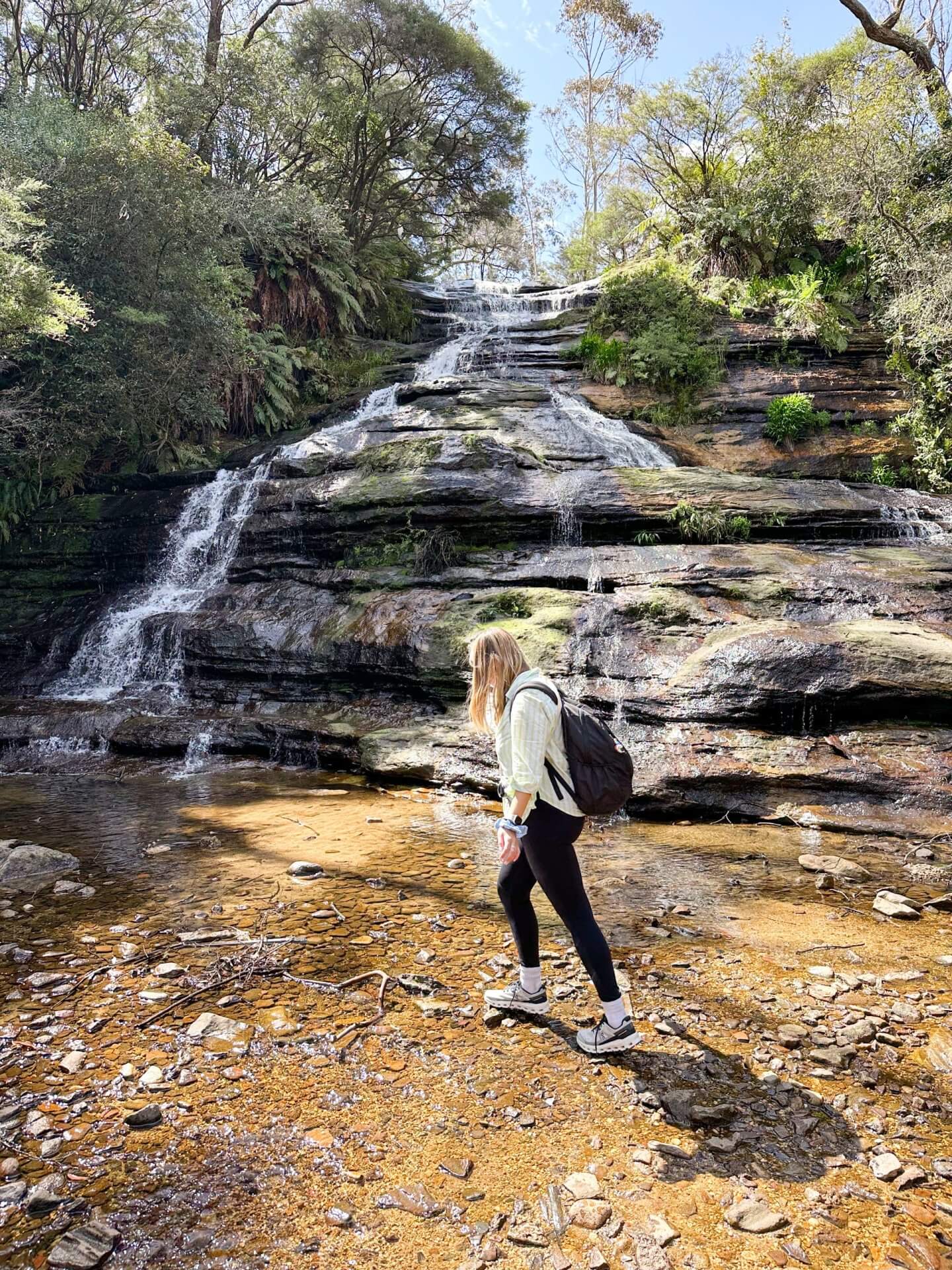 image of a woman walking on stepping stones in front of Katoomba Cascades on a Blue Mountains trip from Sydney