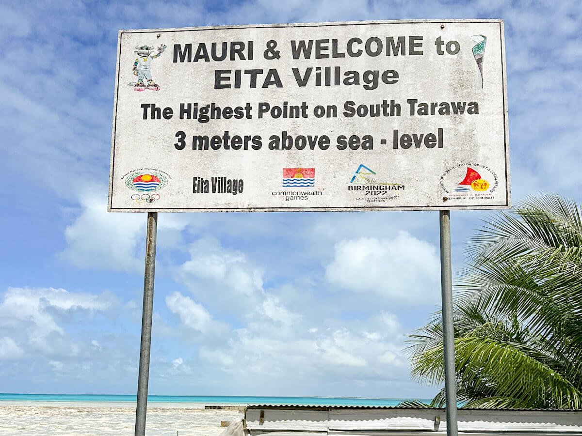 A large sign in Eita Village, South Tarawa, Kiribati, welcomes visitors and states it's "The Highest Point on South Tarawa - 3 meters above sea-level," with the ocean visible in the background.