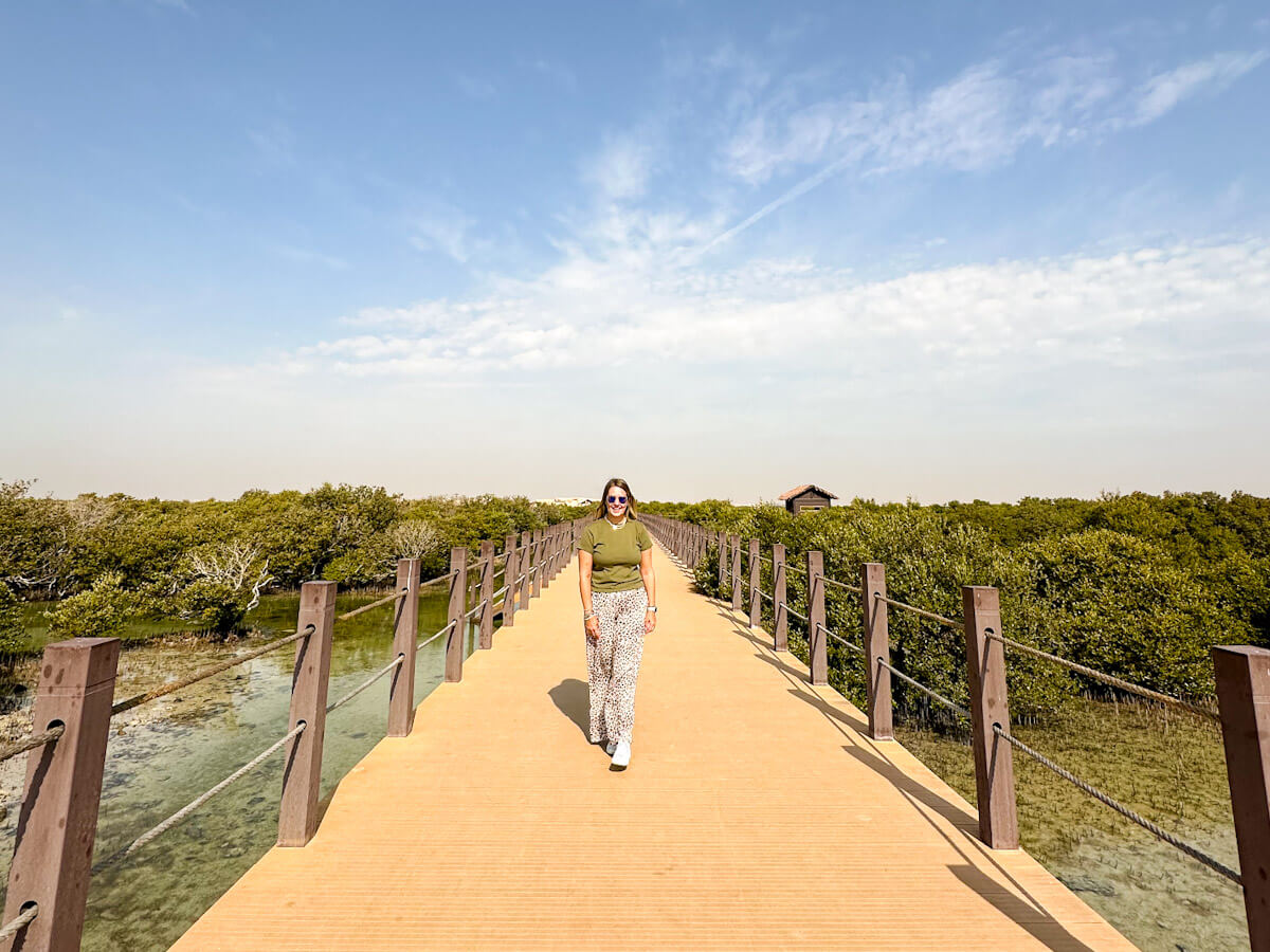 Charlie walks along a wooden boardwalk surrounded by lush green mangroves and clear shallow water at Al Thakira Mangroves in Qatar, under a bright blue sky with scattered clouds.