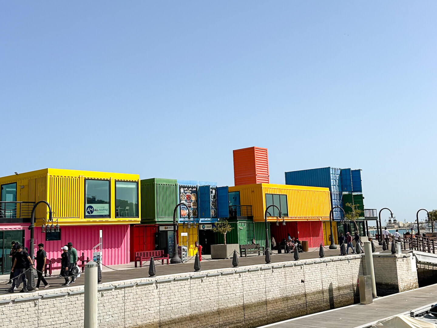 shipping containers painted in bright colours and converted into cafes against a blue sky and sitting next to water at Box Park in Doha