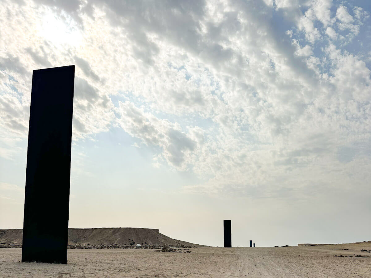 iew of Richard Serra's 'East-West/West-East' art installation in the Qatari desert, featuring four tall, dark steel monoliths standing in a line across a flat, sandy landscape under a cloudy sky