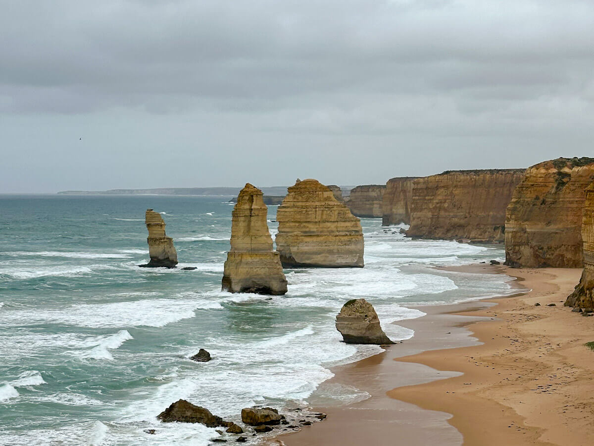Limestone stacks of the Twelve Apostles rise from the sea near a sandy beach and cliffs under a cloudy sky, along the Great Ocean Road