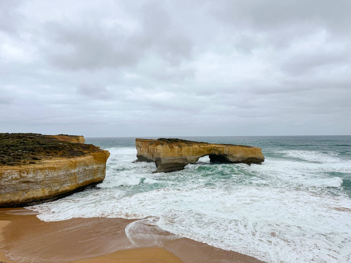 Dramatic waves are crashing against London Bridge on the Great Ocean Road. The sky is overcast and the waves are large