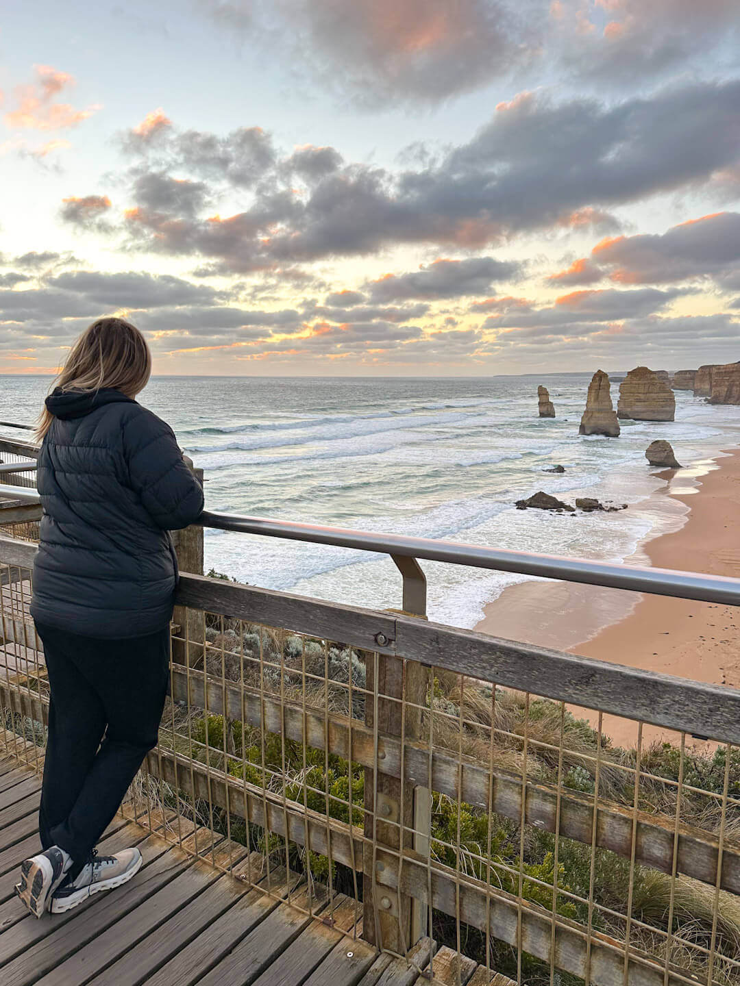 Charlie is leaning on a fence with her legs crossed behind her. She is looking out over the Twelve Apostles at sunset