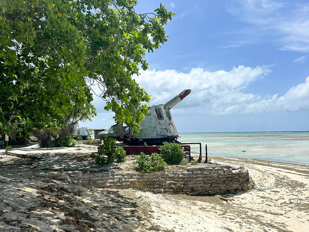 Weathered WWII Japanese gun on a sandy beach in Betio, Kiribati, facing a calm, light blue ocean under a partly cloudy sky.