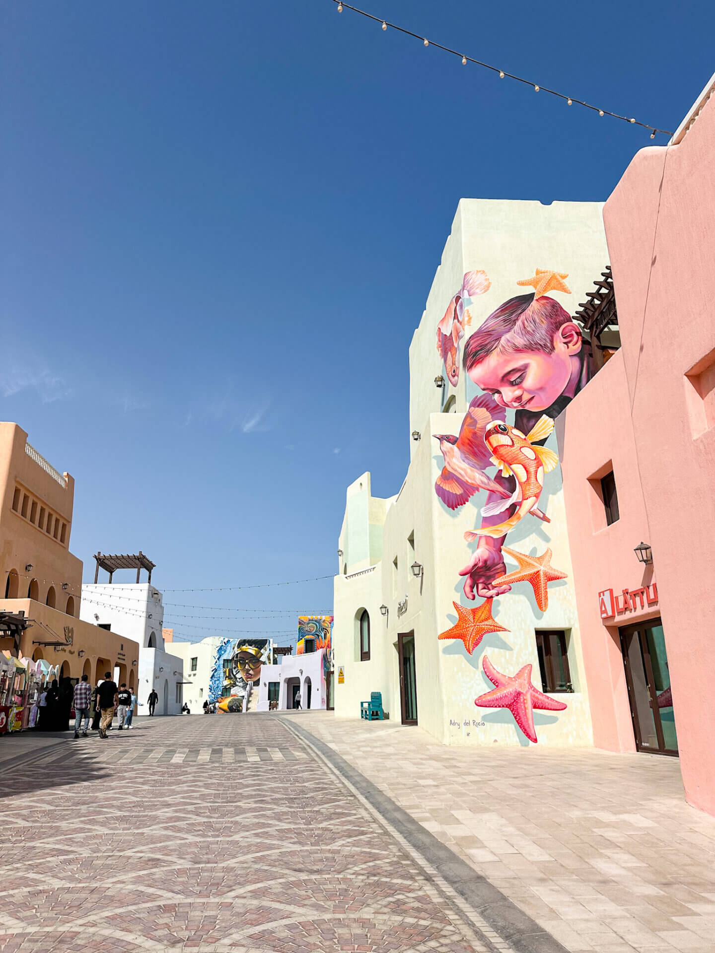 A pink and orange mural of a boy looking down at star fish is painted on a pastel mint building in the Mina District in Doha. A street can be seen in the middle leading to more street art
