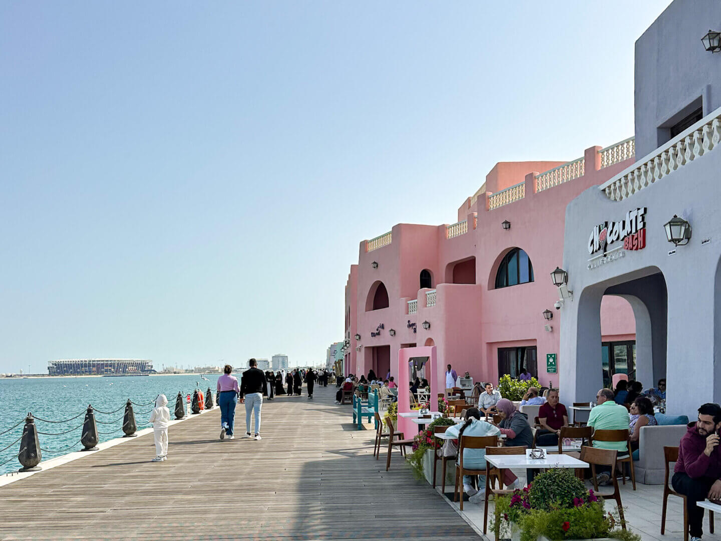 Pastel pink and purple cafes are on the right hand side, with people sitting on outside furniture enjoying lunch. In the middle is a boardwalk and on the left the sea. Mina District which is one of the best places to see in Doha.
