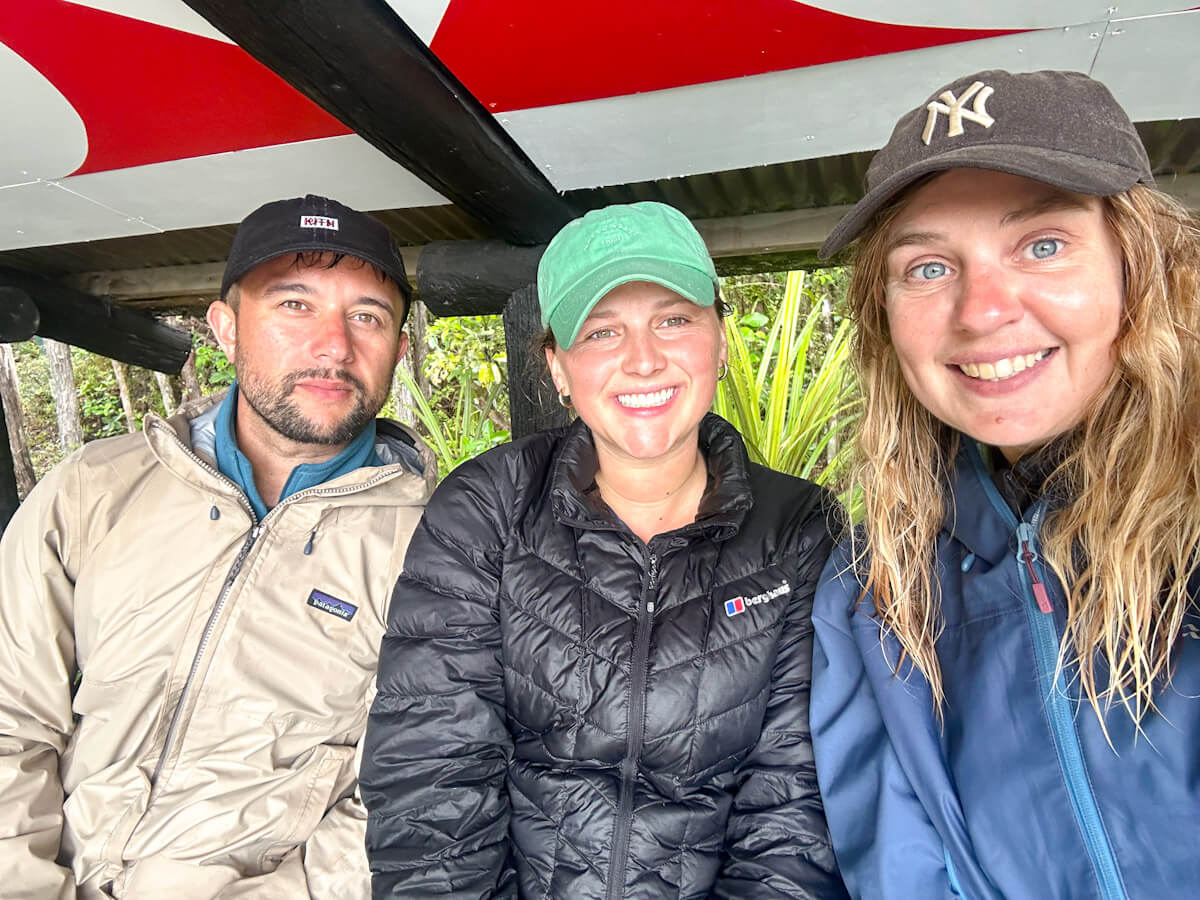 Three people are sitting side by side having completed the Tongariro Crossing in New Zealand. The are all wearing coats and hats and are damp