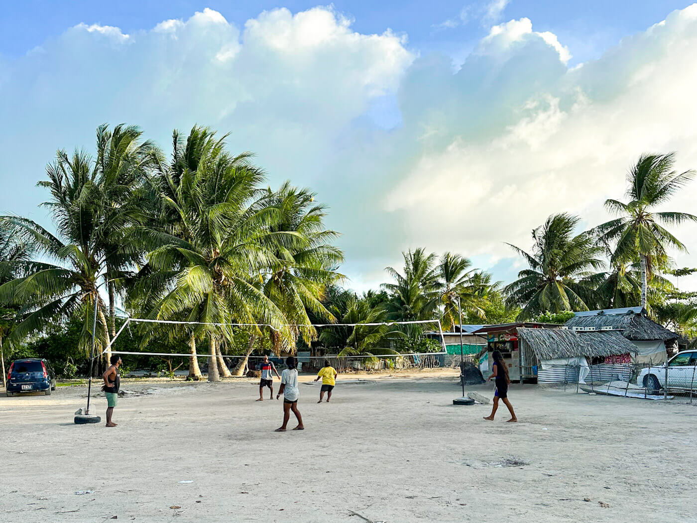 children are playing volleyball in a village in North Tarawa, Kiribati. Palm trees and behind the net and buildings with weaved roofed on the right