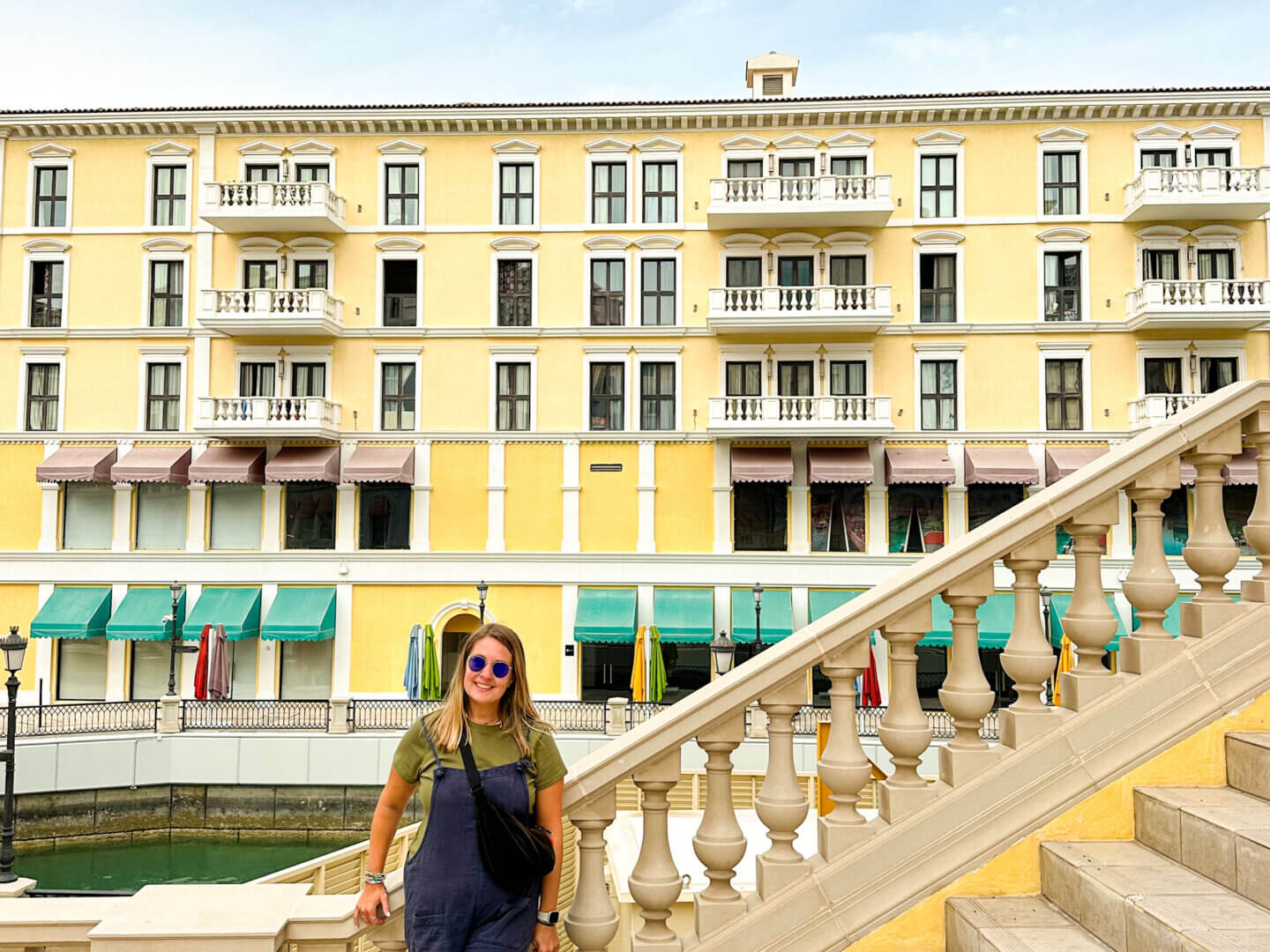 Charlie stands on a steps up to a bridge. Behind her is yellow venetian style buildings in yellow with white windows