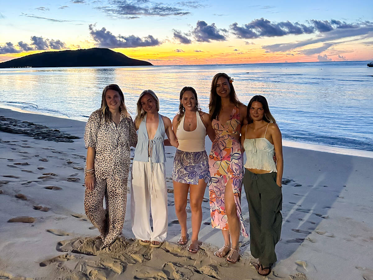 Five people are standing with their arms around each other on a beach in Fiji at sunset. The sky behind them is blue, yellow and orange