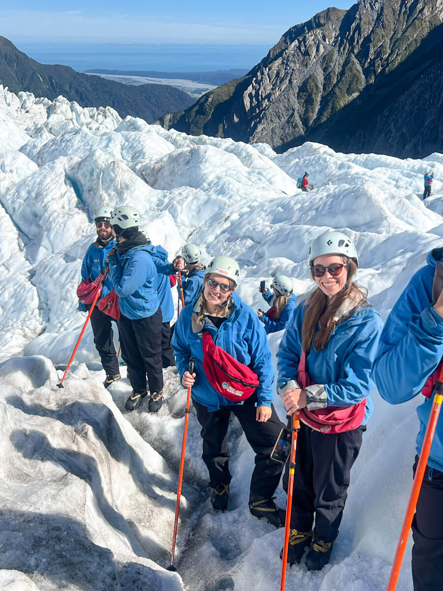 A group of people are standing on Franz Josef Glacier. They are wearing thick clothes of blue jackets, black trousers, white helmet and carrying red poles with crampons on. The sky is blue