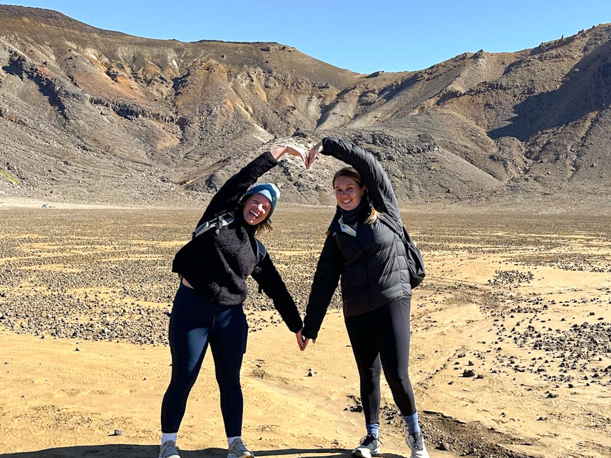 two women are standing in front of the volcanic landscape of the Tongariro Crossing. They are leaning towards each other and making a heart shape with their arms.