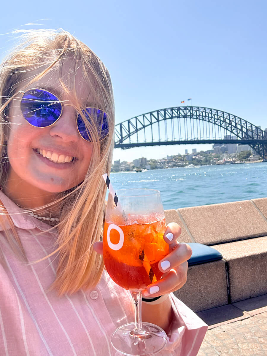 Charlie sitting in front of the Harbour Bridge in Sydney. She is wearing a pink shirt, smiling and holding an aperol