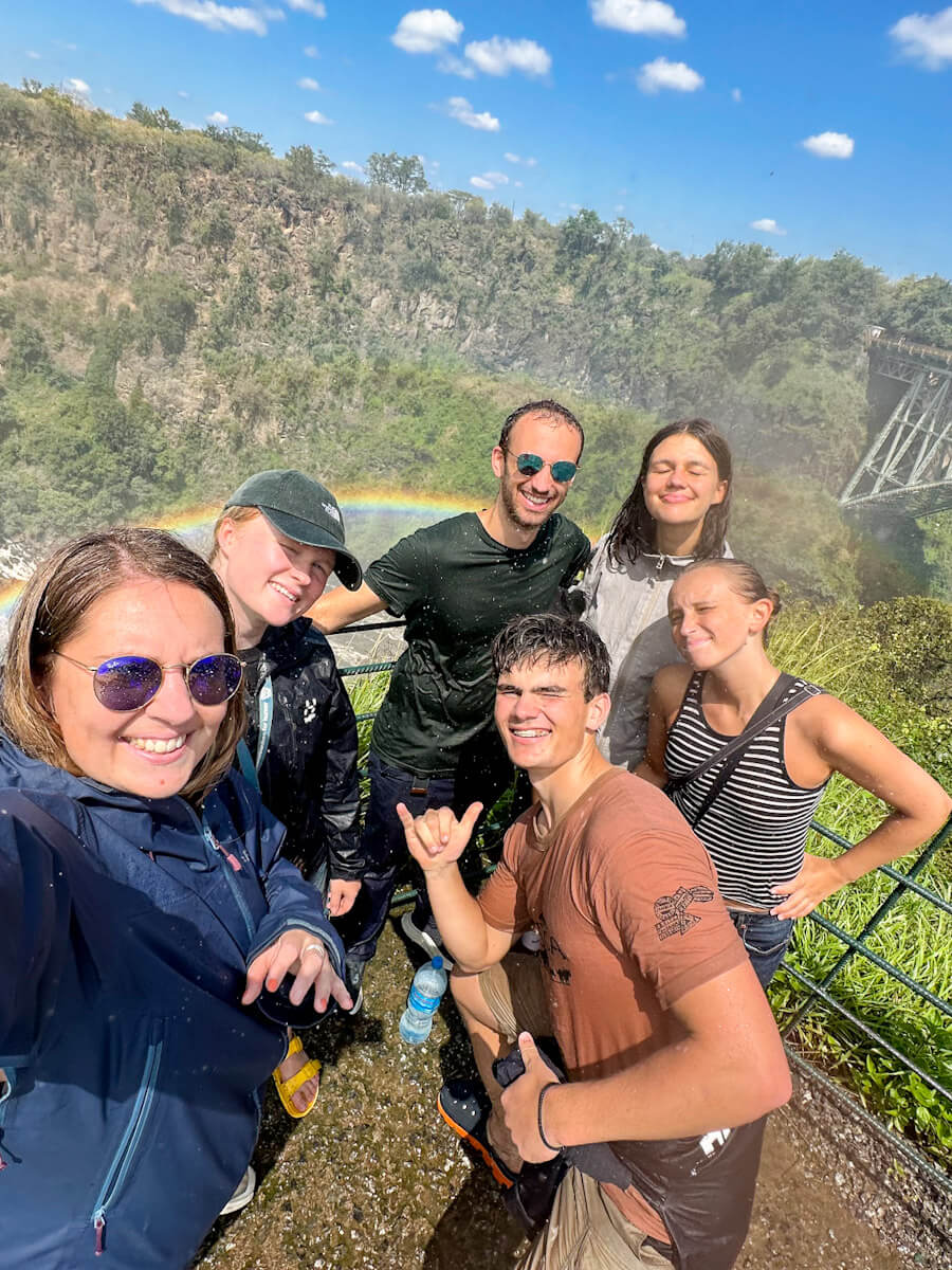 A group of people are standing in front of a rainbow at Victoria Falls, Zimbabwe. They are very wet from the spray of the waterfall