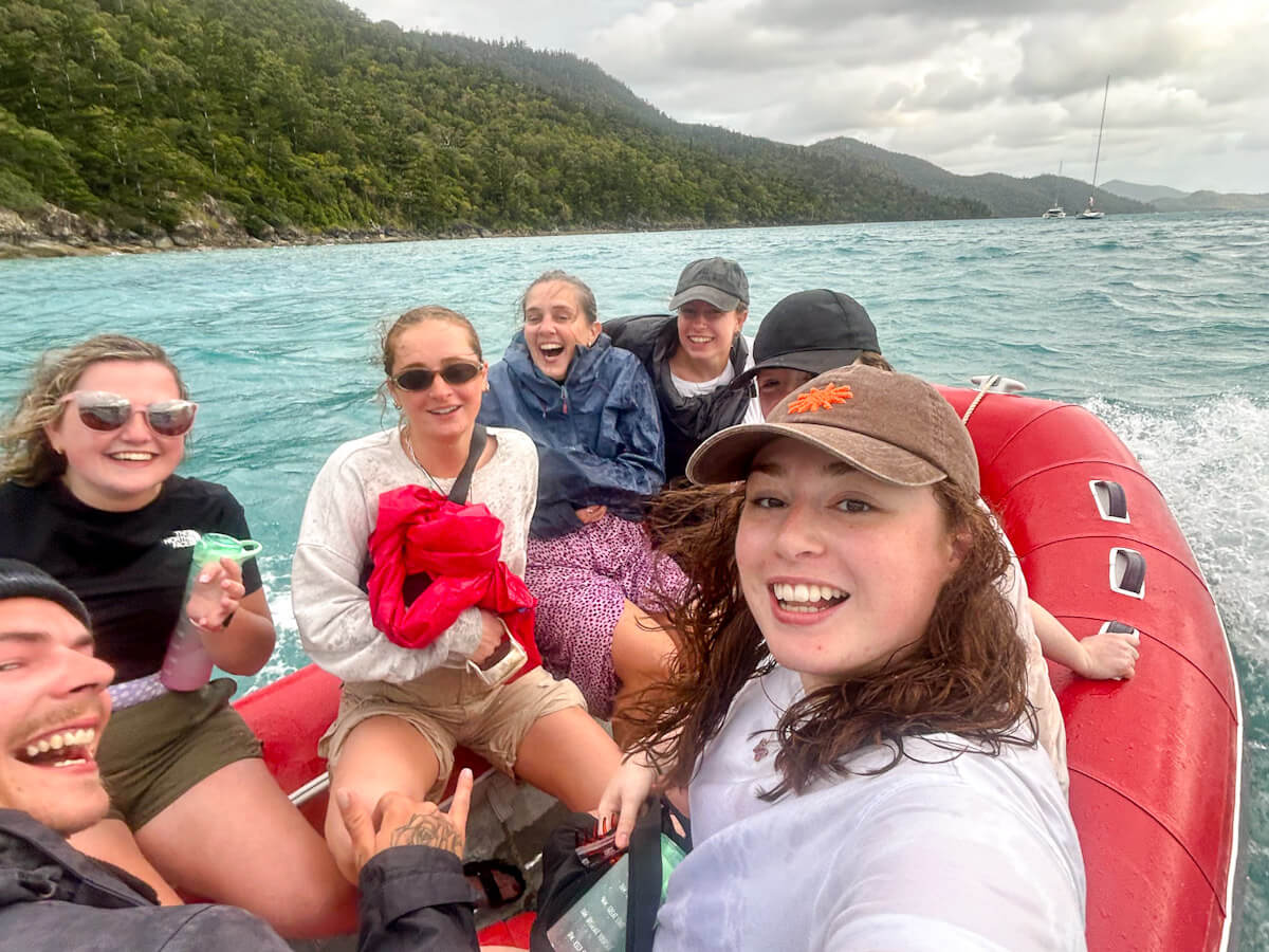A group of people are sitting on a red rib boat. Water is spraying over them and they are laughing as they are so wet