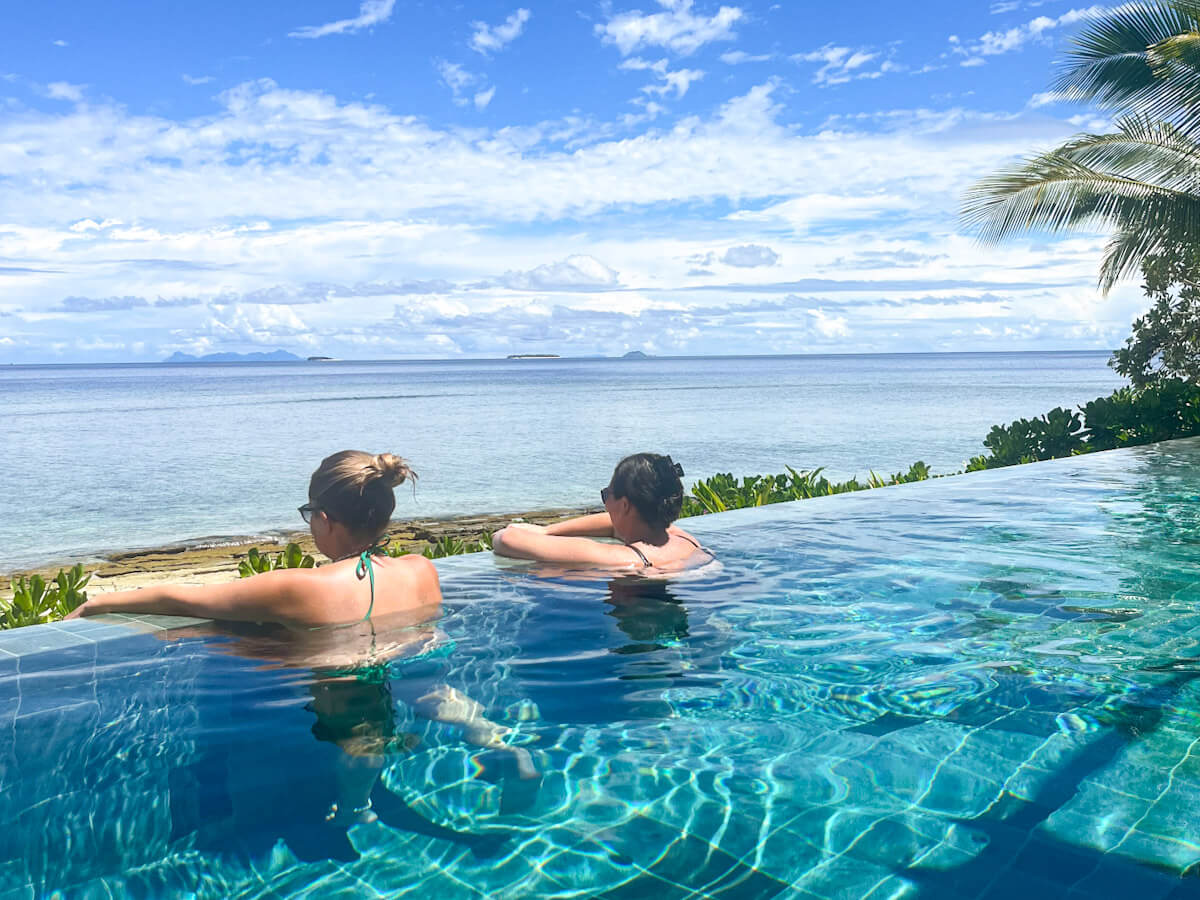 two people are in an infinity swimming pool. They have their backs to the camera, arms over the edge and are looking out over the sea in Fiji