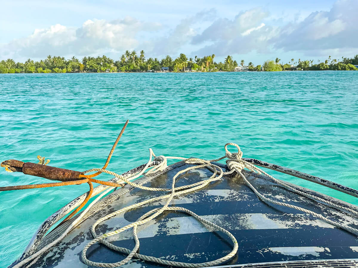 the front of a boat while crossing Sue's Creek in North Tarawa, Kiribati. The sea is a bright turquoise and palm trees can be seen in the distance.