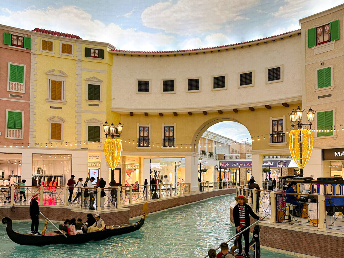 two gondolas and punters on an indoor canal in Villaggio Mall, Doha. There is an arch that the canal goes through and shops lining either side underneath a painted blue sky
