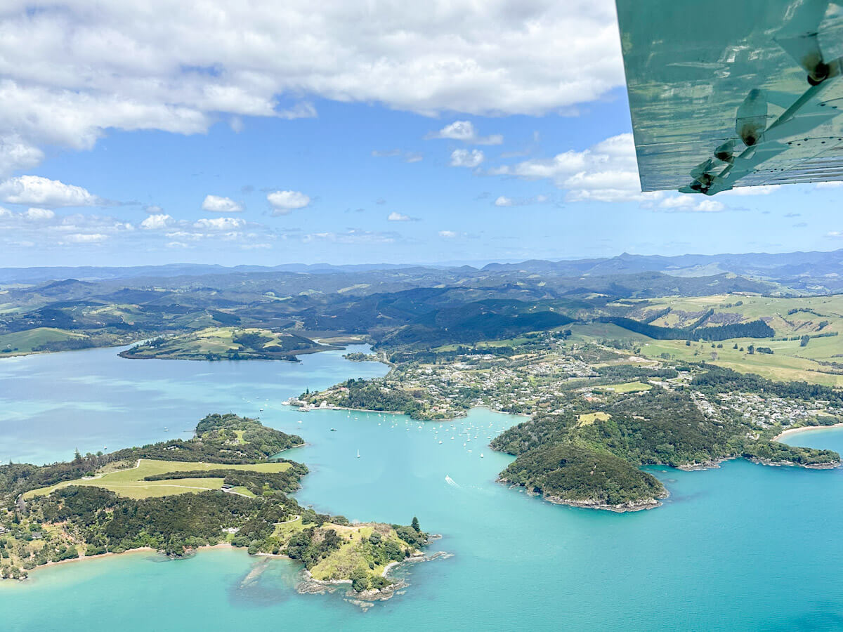 An expansive aerial view of the stunning Bay of Islands, showcasing numerous lush green islands scattered across vibrant turquoise waters dotted with boats. The distant coastline reveals rolling hills and scattered settlements, providing a breathtaking perspective of this popular New Zealand destination.