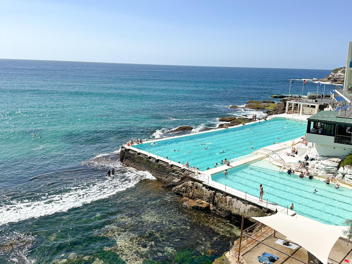An eye-level shot of Bondi Icebergs Pool with swimmers, nestled between the turquoise ocean and a building complex, under a clear blue sky.