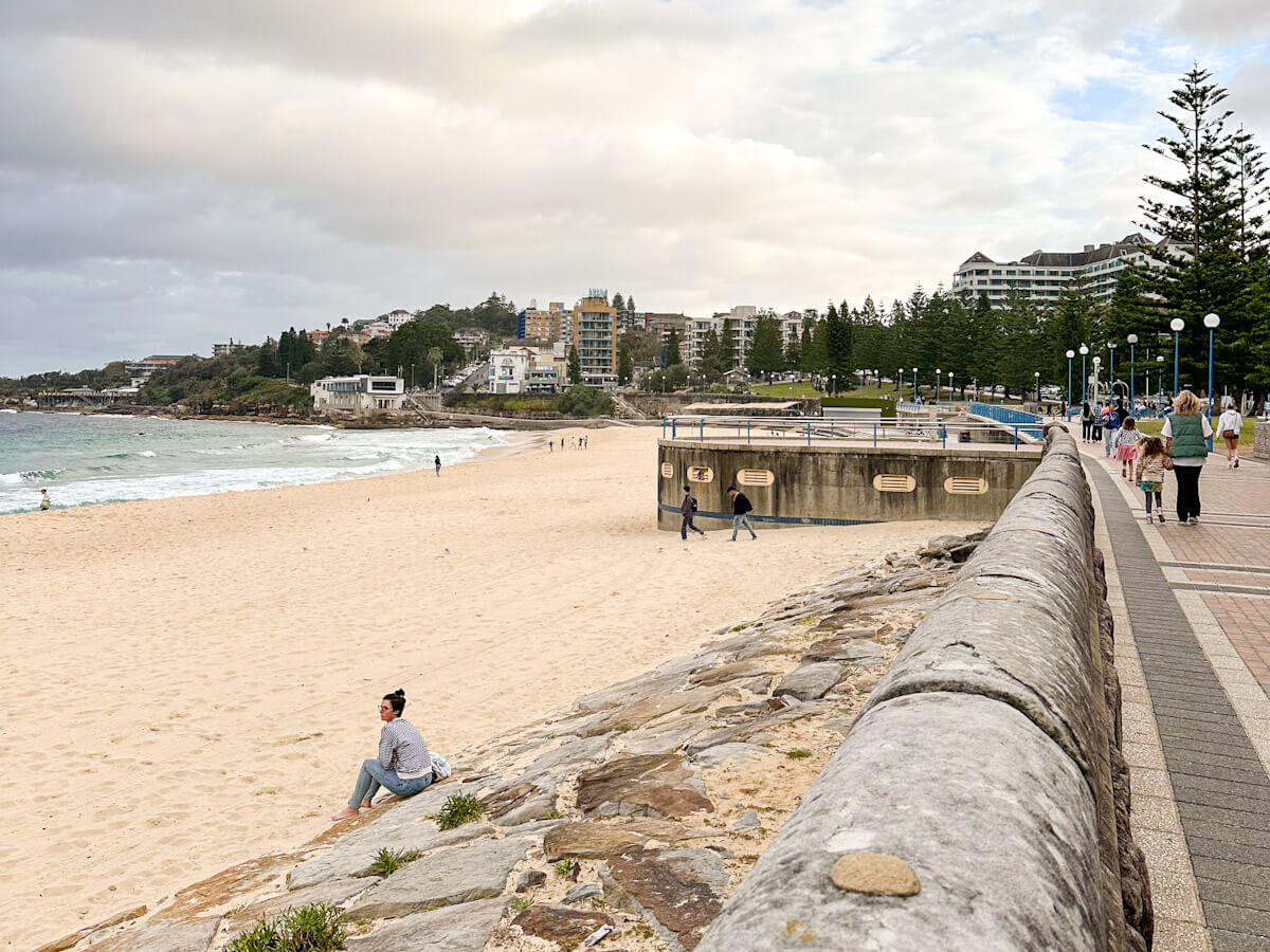 A wide sandy beach bordered by a promenade with people walking, facing the ocean, with buildings and trees on the distant hills under an overcast sky.