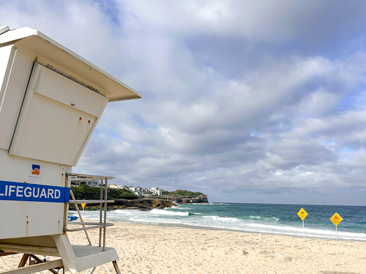 A white lifeguard tower stands on Bronte Beach, a wide sandy beach, facing the ocean with breaking waves and distant cliffs under a cloudy sky.