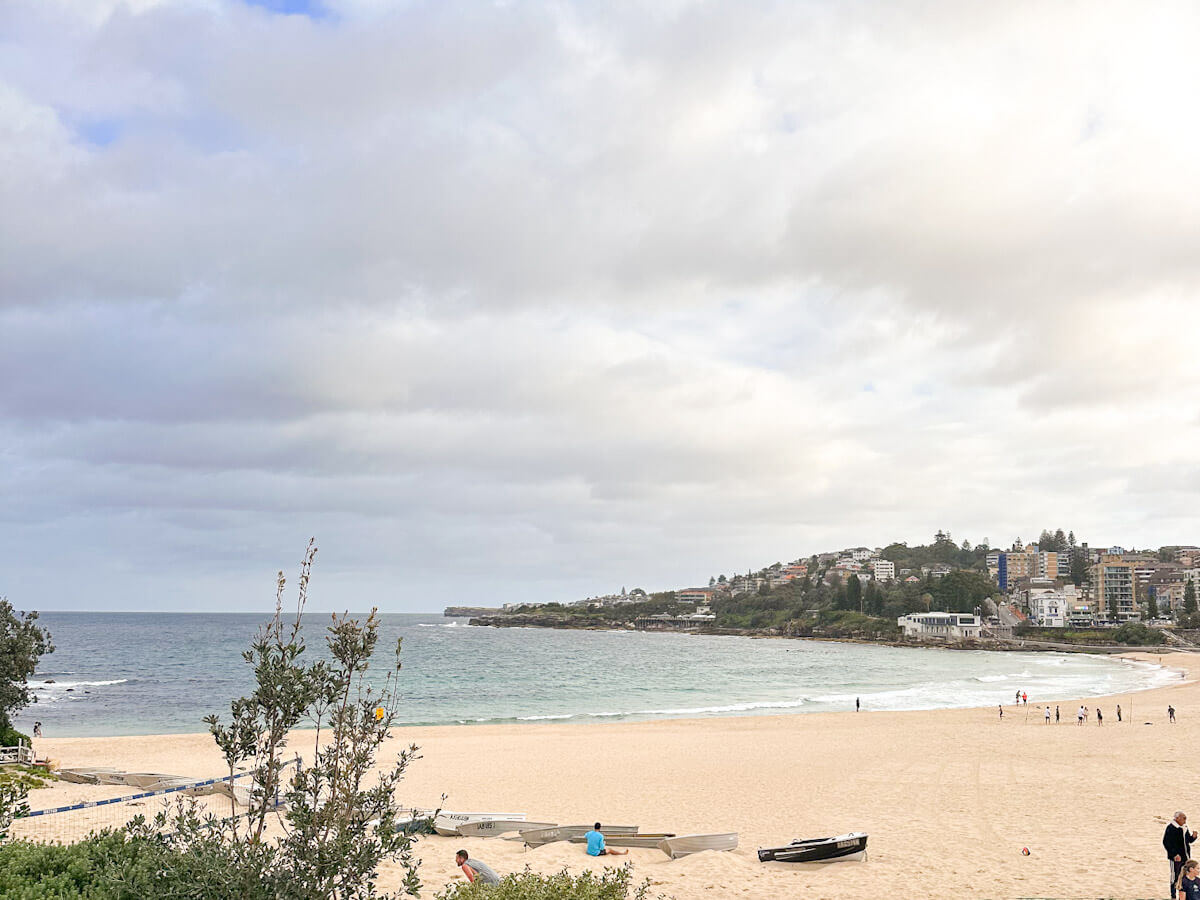 Coogee Beach on an overcast day, featuring a wide sandy expanse with scattered people and boats, facing the ocean, with a headland and distant buildings on the horizon.