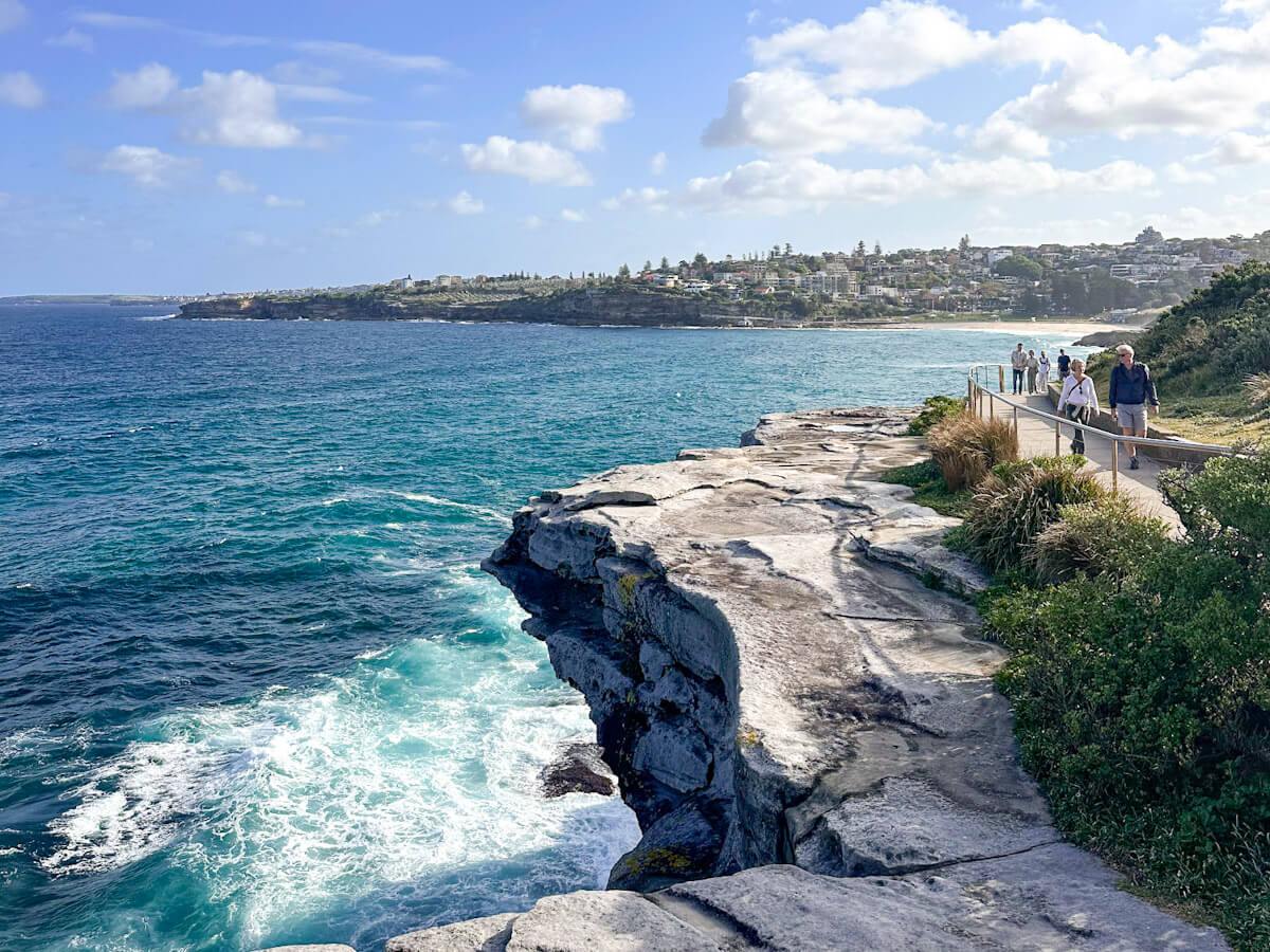 A coastal walking path on a cliff edge overlooking the deep blue ocean with crashing waves, with people walking in the distance towards a sandy beach and town under a partly cloudy sky.