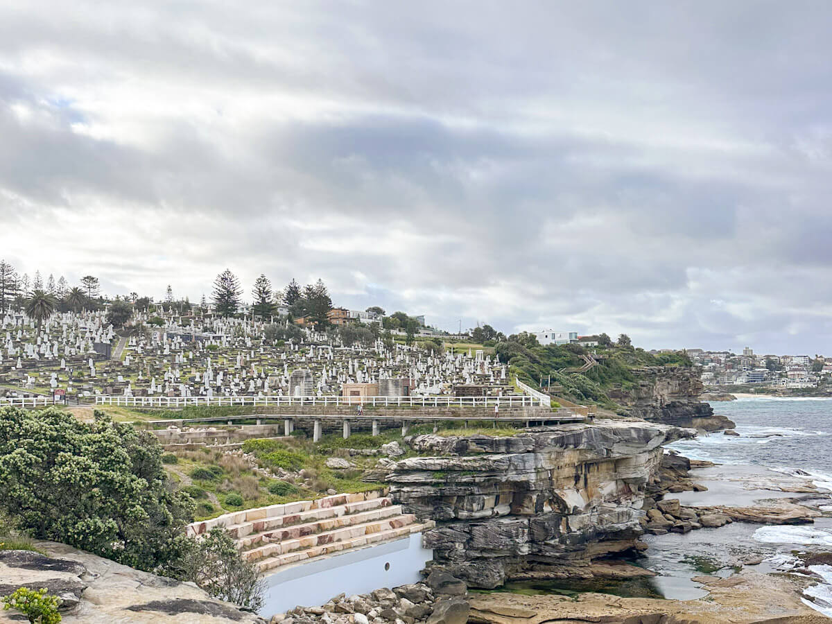Waverley Cemetery, a large hillside cemetery filled with white tombstones, overlooks a rocky coastline with a natural swimming pool and the ocean under a cloudy sky.