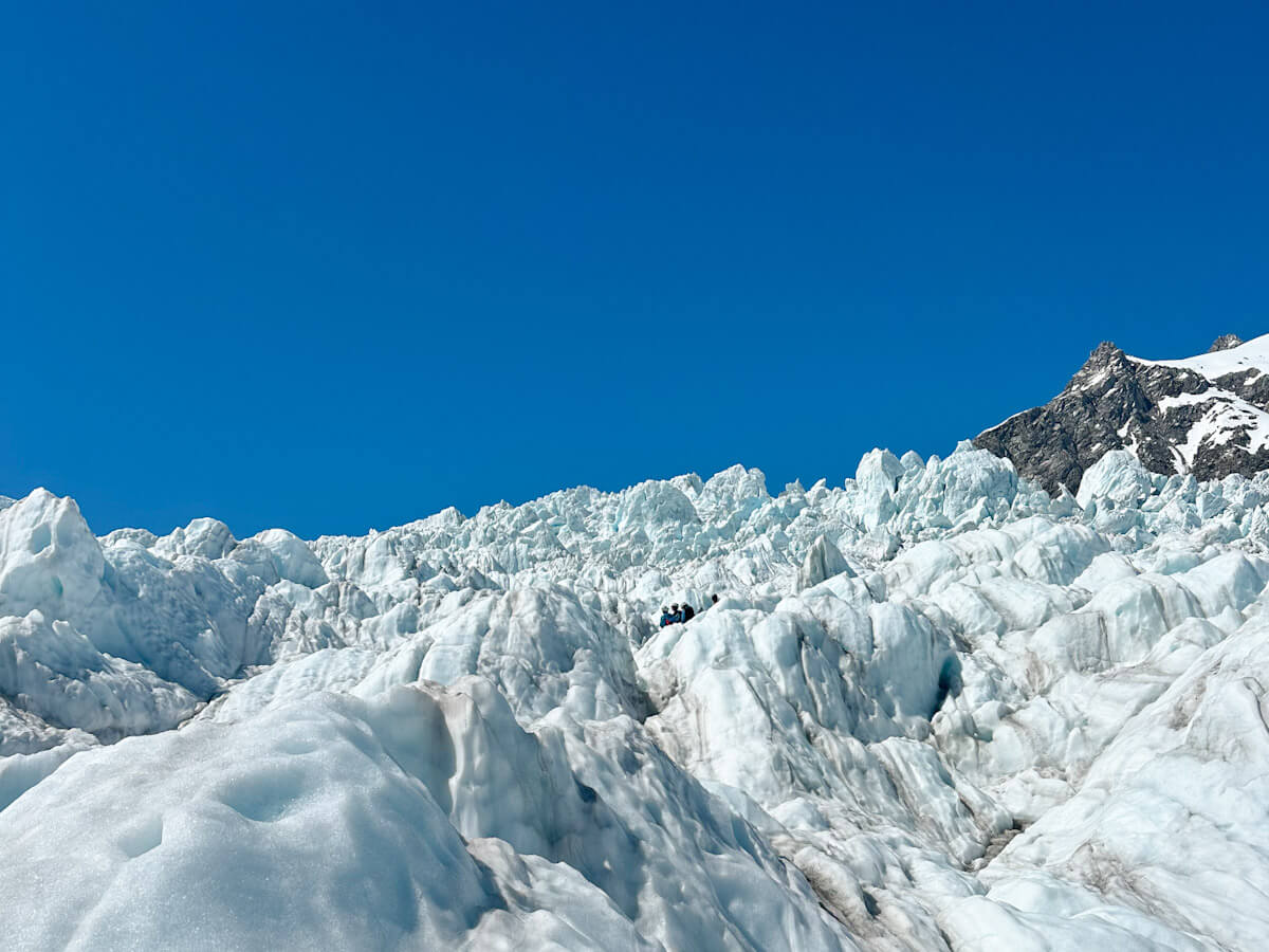 A breathtaking close-up of the Franz Josef Glacier, showcasing its impressive, rugged ice formations under a clear, bright blue sky. Small figures of people can be seen in the distance on the glacier, highlighting its immense scale.
