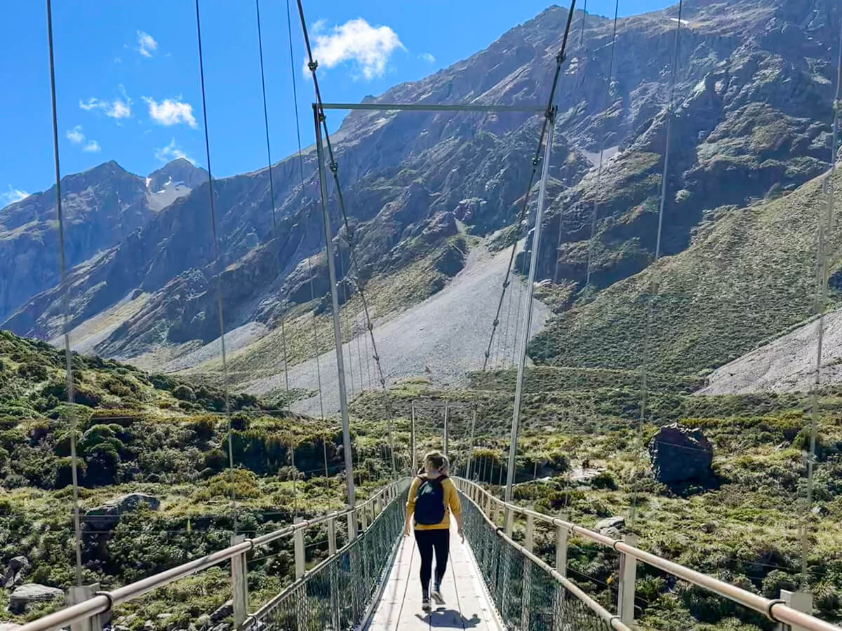 Charlie walks across a suspension bridge along the Hooker Valley Track, with dramatic, rugged mountains rising majestically in the background under a bright blue sky. The scene captures the adventurous spirit of hiking in the Aoraki/Mount Cook region.