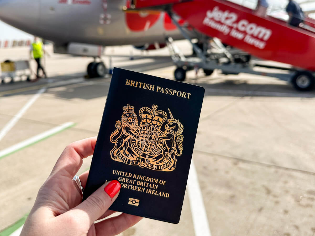 A hand with red painted fingernails holds open a black British passport with a gold crest, showing the words "BRITISH PASSPORT" and "UNITED KINGDOM OF GREAT BRITAIN NORTHERN IRELAND." In the blurred background, a red Jet2.com airplane and airport staff are visible on the tarmac.