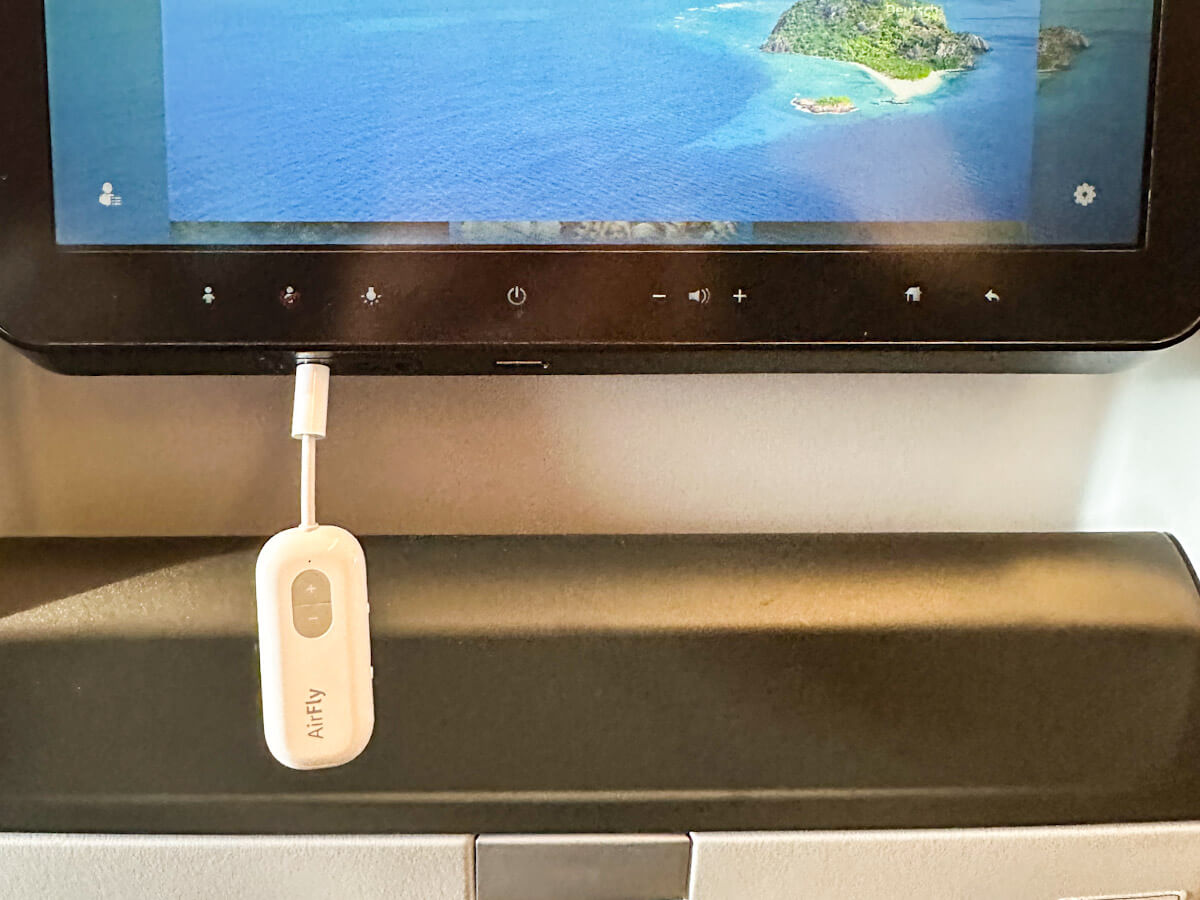 A white "AirFly" device with a short cable is plugged into the audio jack of an airplane entertainment screen. The screen displays a map with an island surrounded by blue water.