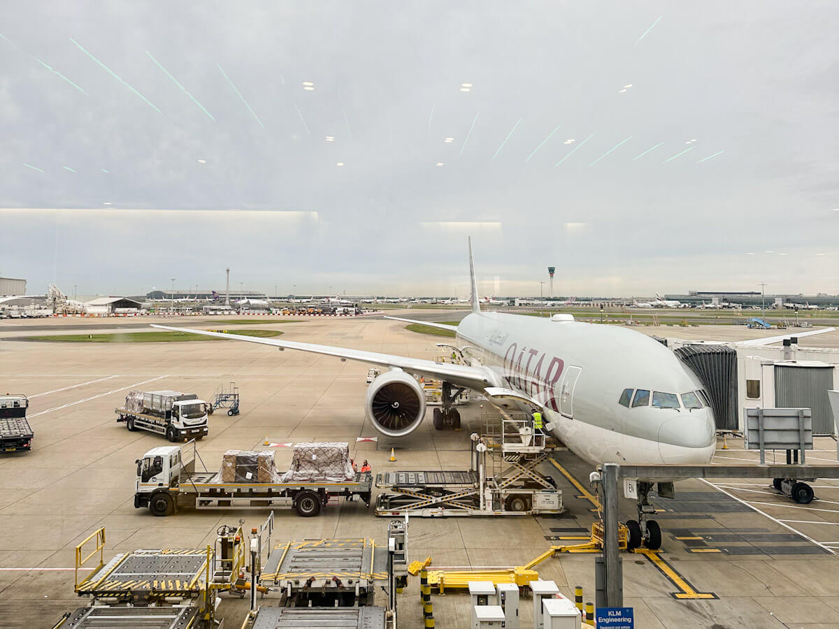 A large white Qatar Airways plane is parked at a gate at Heathrow Airport under an overcast sky. Ground crew and vehicles are around the plane, loading cargo. Other airport buildings and planes are visible in the background.