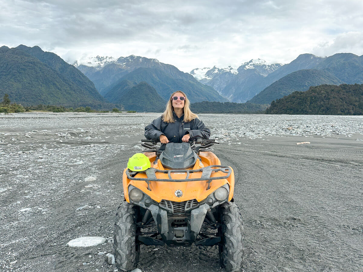 Charlie is smiling brightly while sitting on a bright yellow quad bike in a wide, rocky riverbed near Franz Josef. In the background, majestic mountains with snow-capped peaks rise under a cloudy sky.