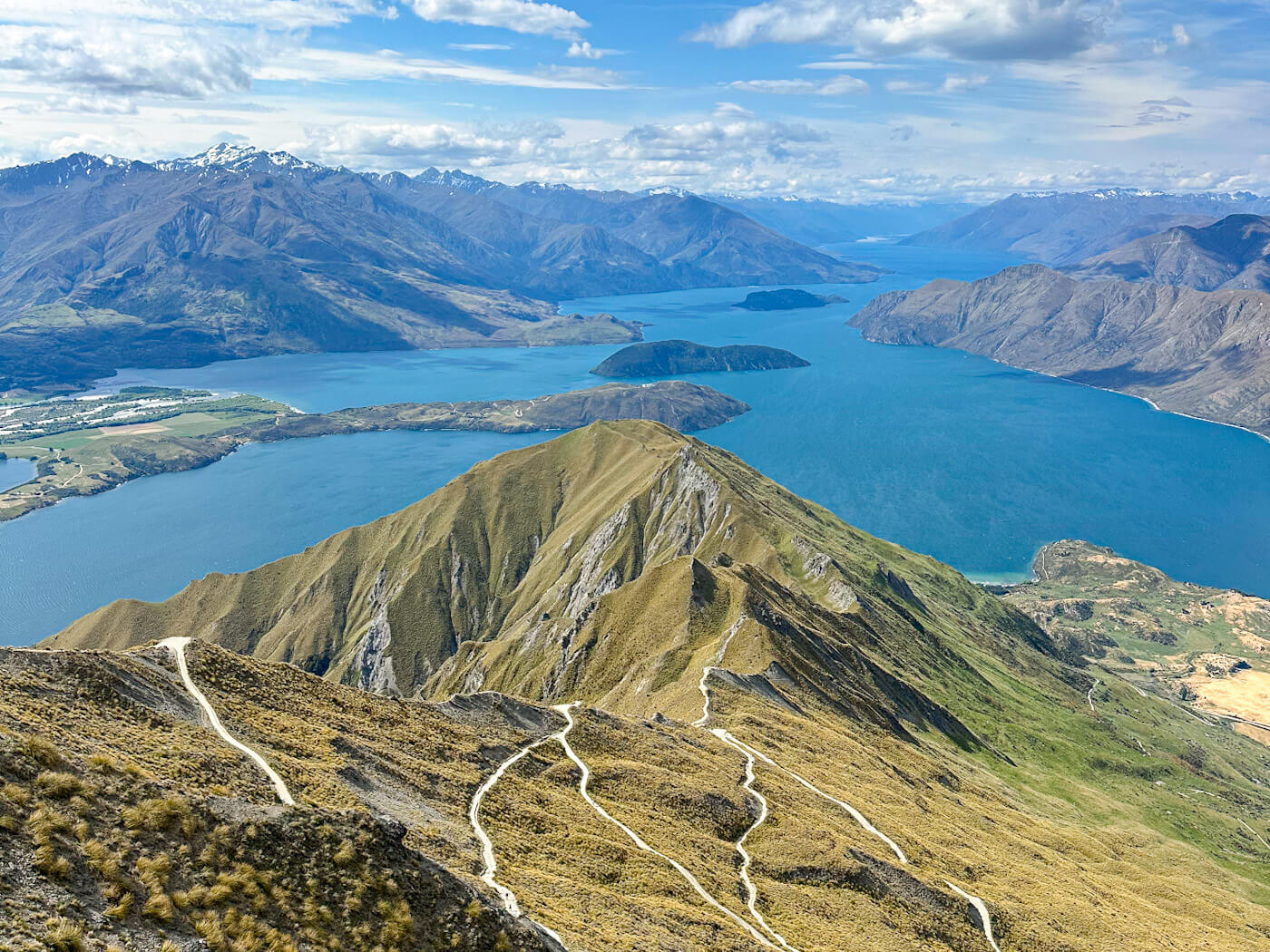 A breathtaking panoramic view from Roy's Peak in Wanaka, New Zealand, showcases winding hiking trails on grassy slopes, leading down to a vast, deep blue lake surrounded by majestic snow-capped mountains under a bright sky with scattered clouds.