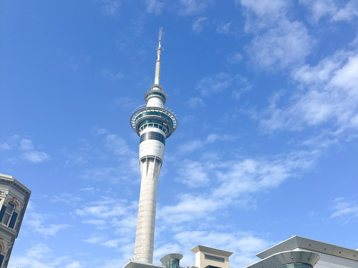 A clear shot of the iconic Sky Tower dominating the Auckland skyline, its slender white structure reaching towards a bright blue sky with wispy white clouds. The bottom part of the image shows architectural details of surrounding buildings.
