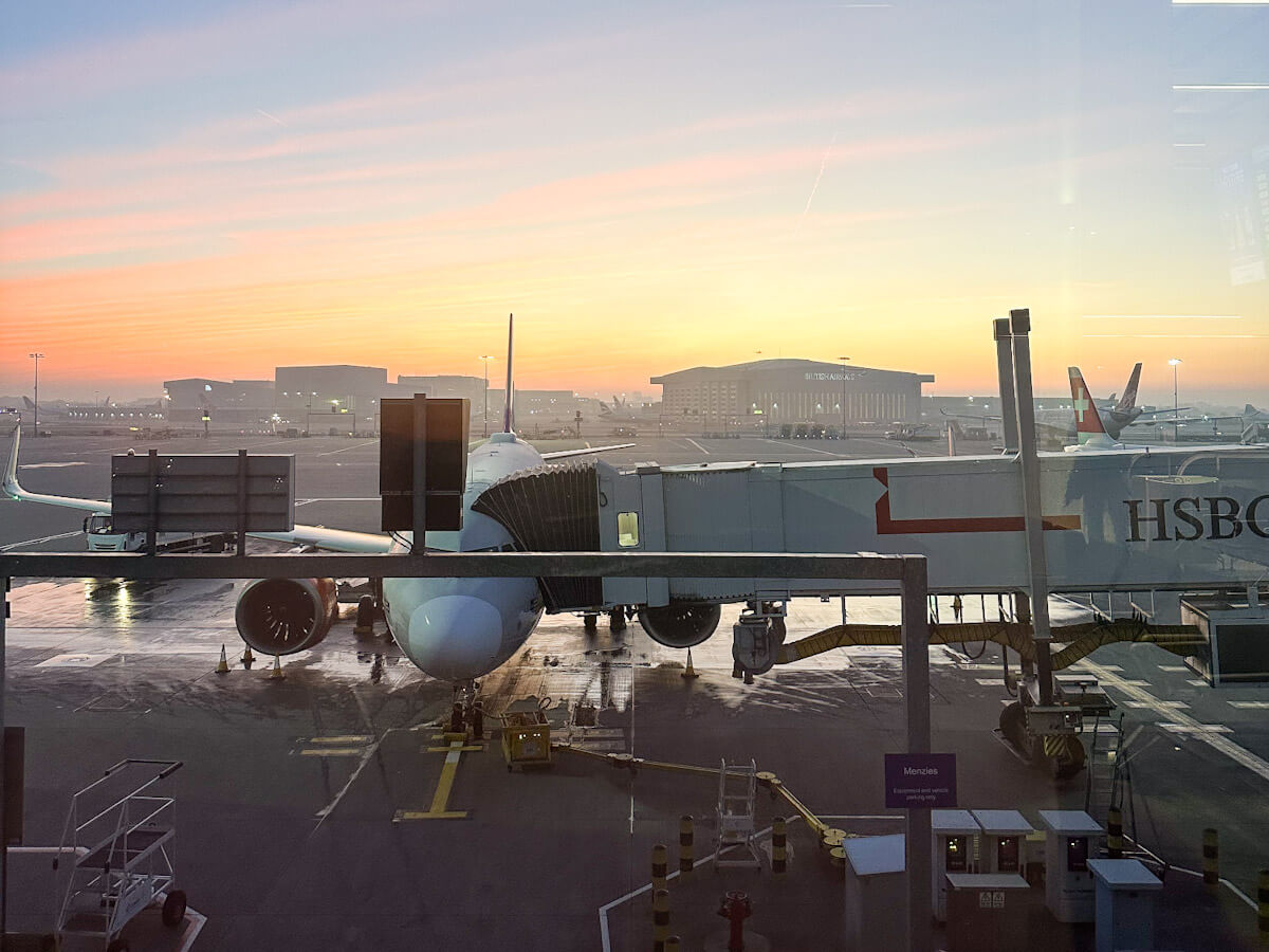 A Swiss Airlines plane is connected to a jet bridge at an airport gate during a vibrant orange and pink sunset or sunrise. The tarmac is wet, reflecting the light. Other airport buildings and planes are visible in the foggy distance.