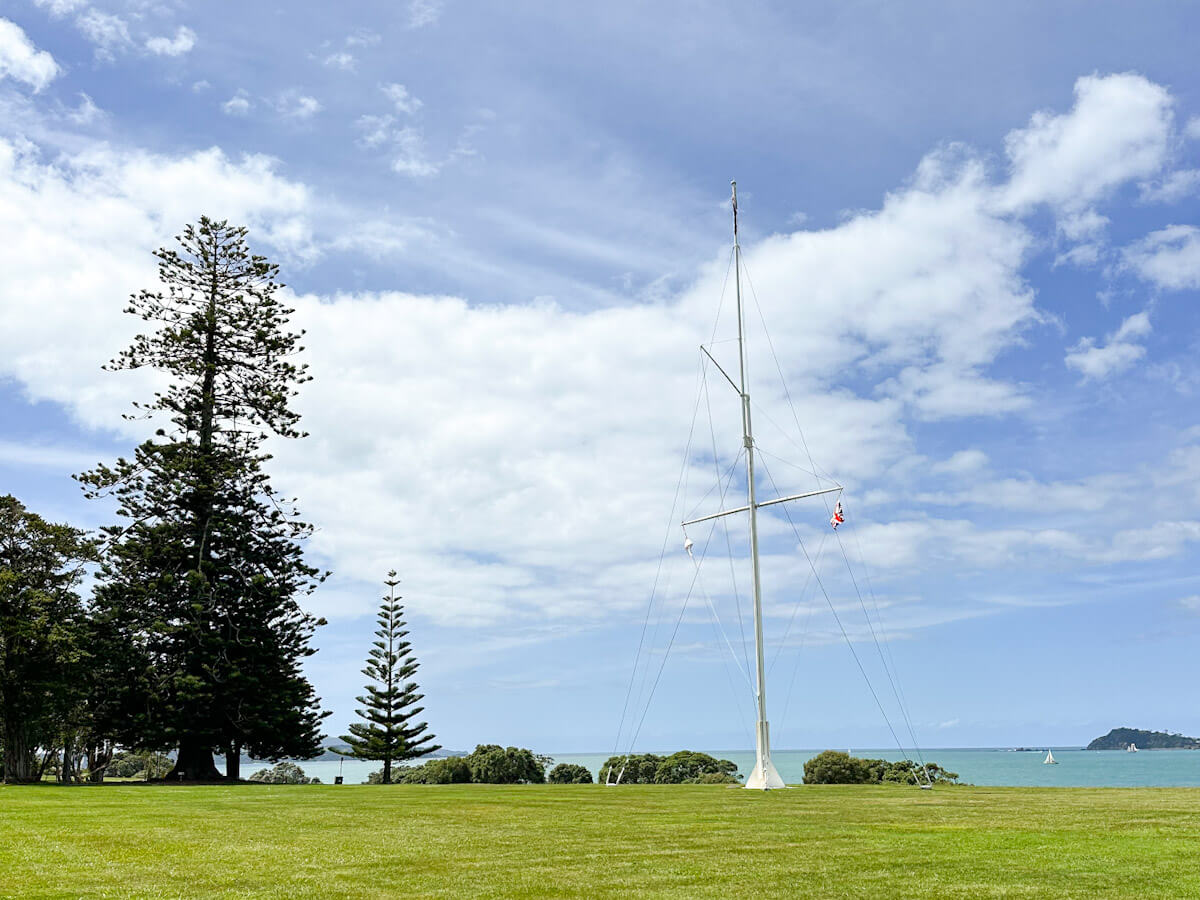 A wide shot of the expansive green lawns at the Waitangi Treaty Grounds, featuring tall trees on the left and a prominent flagpole in the center. In the background, the serene blue water of the bay is visible under a cloudy sky, with sailboats in the distance.