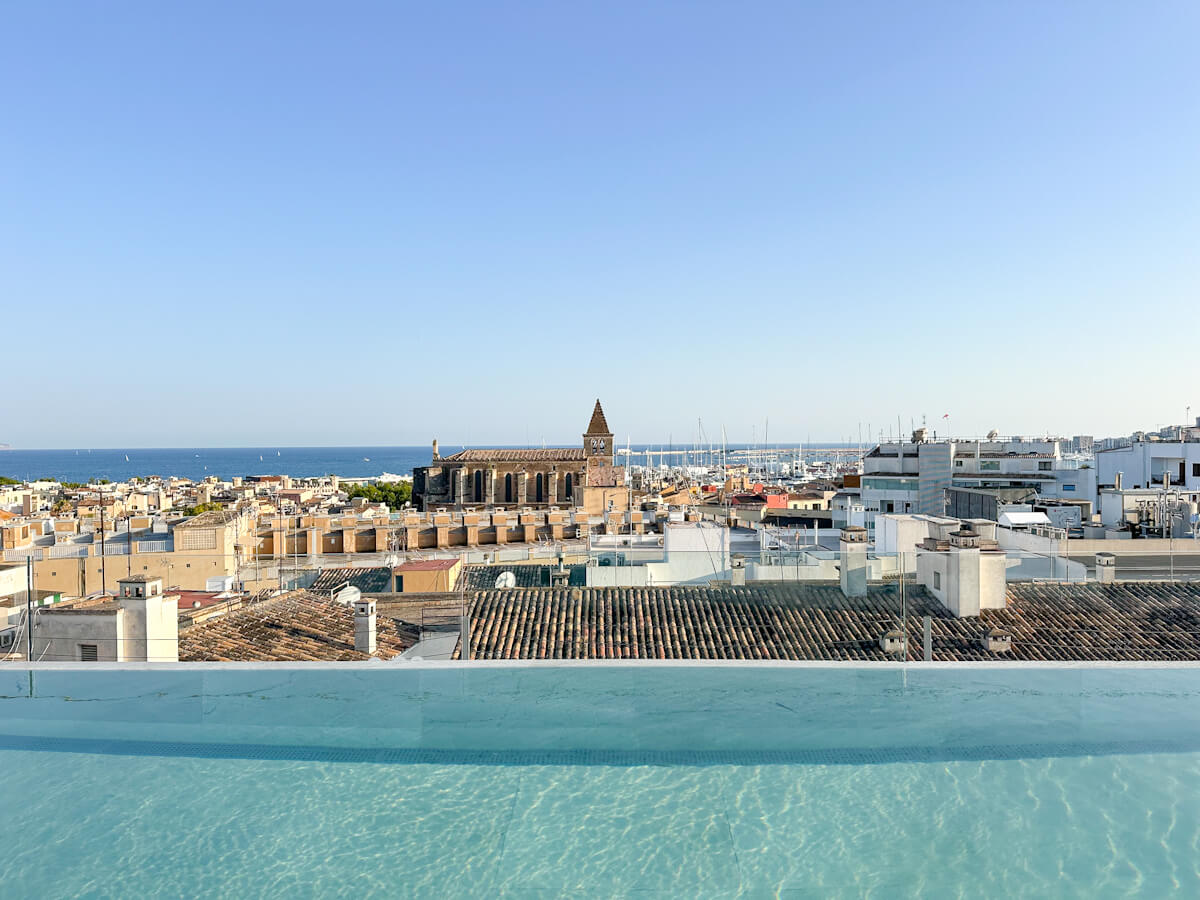 From the Nakar Hotel rooftop bar in Palma de Mallorca, an infinity pool in the foreground offers a panoramic vista of the city, with the sea, boats, and a historic steeple-topped building under a clear sky.