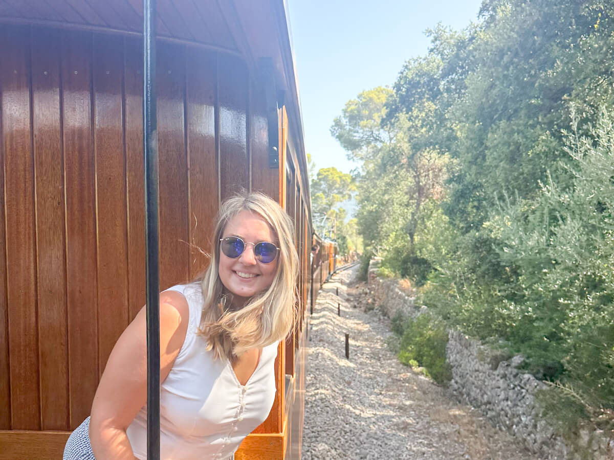 Charlie is in sunglasses smiling brightly, leaning out of a vintage wooden Soller train window in Palma de Mallorca as it chugs alongside a rustic stone wall and lush green trees.