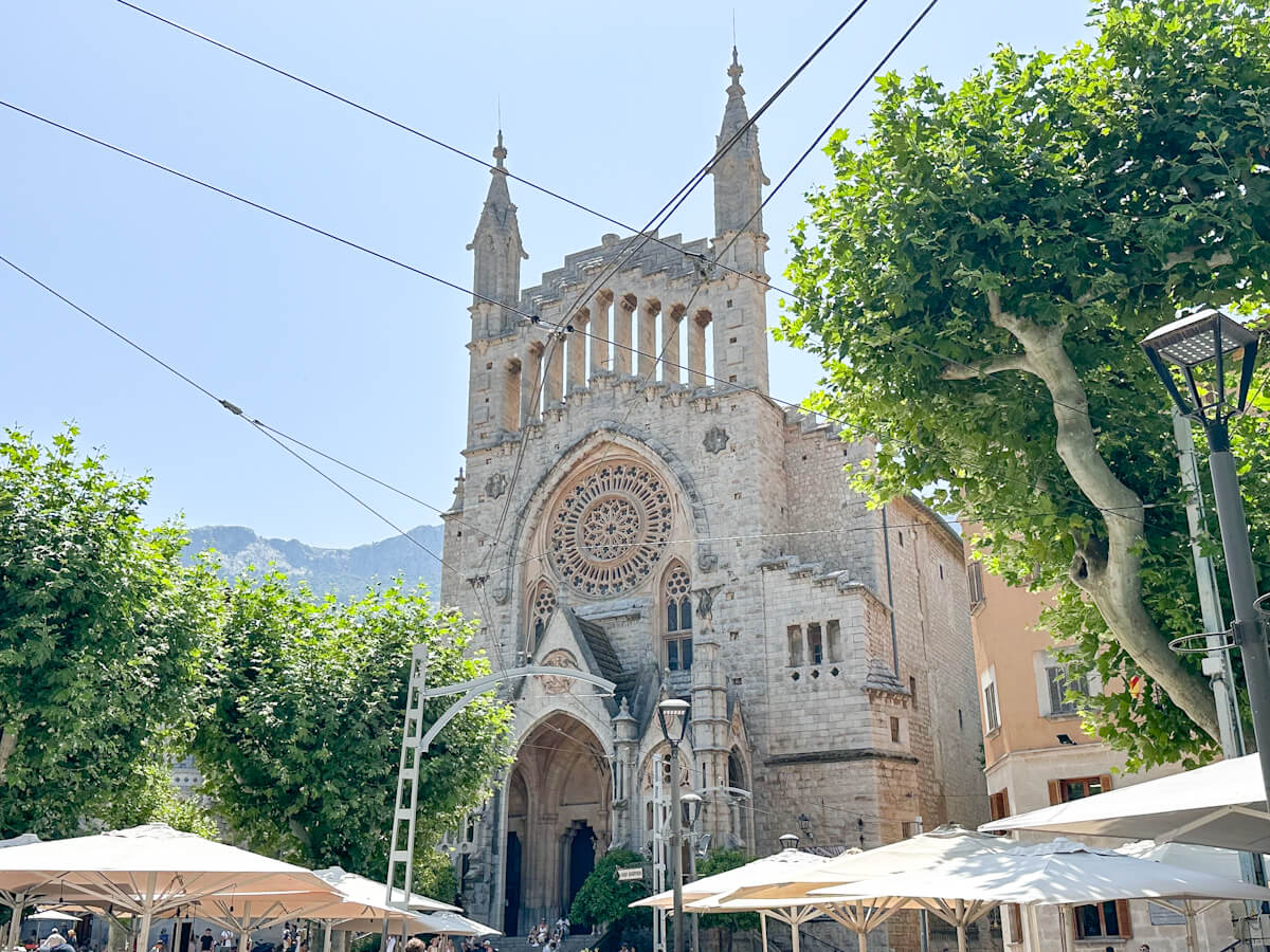 In Soller, Mallorca, the grand, ornate stone facade of a church, complete with spires and a stunning rose window, is framed by vibrant green trees and the welcoming umbrellas of outdoor cafes, all bathed in bright sunlight.