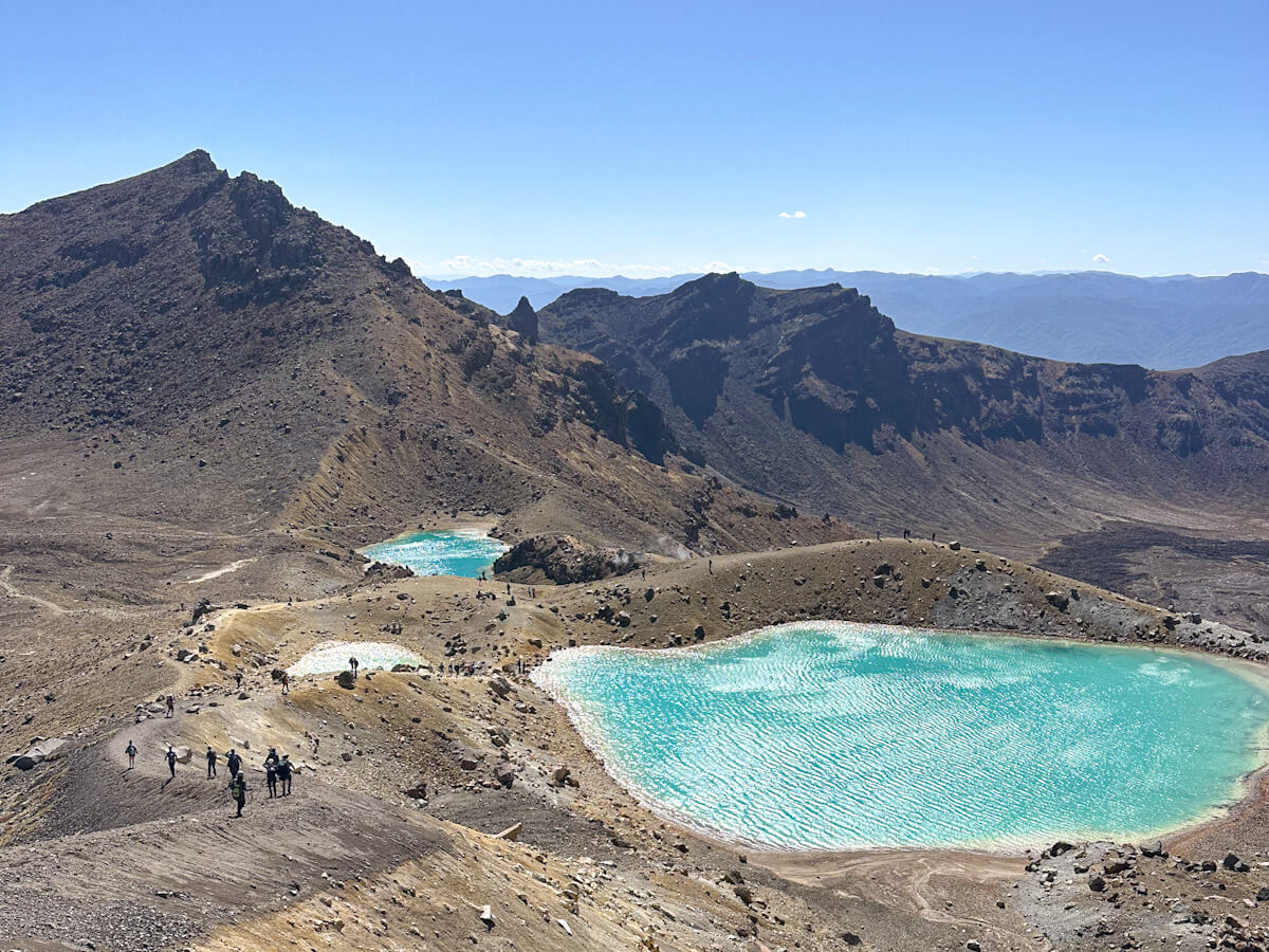 Panoramic view of the stunning turquoise Emerald Lakes and other volcanic features along the Tongariro Alpine Crossing, with hikers enjoying the clear day in New Zealand.