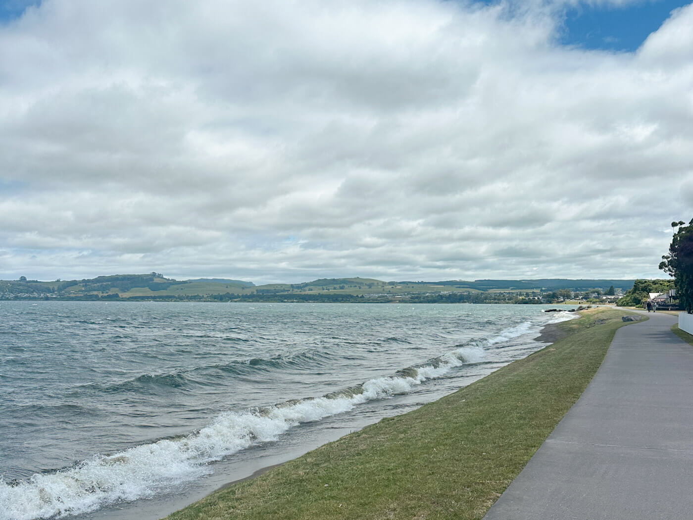 A scenic view of Lake Taupo's shoreline with gentle waves and a paved walking path, perfect for a stroll during a visit to New Zealand's North Island.