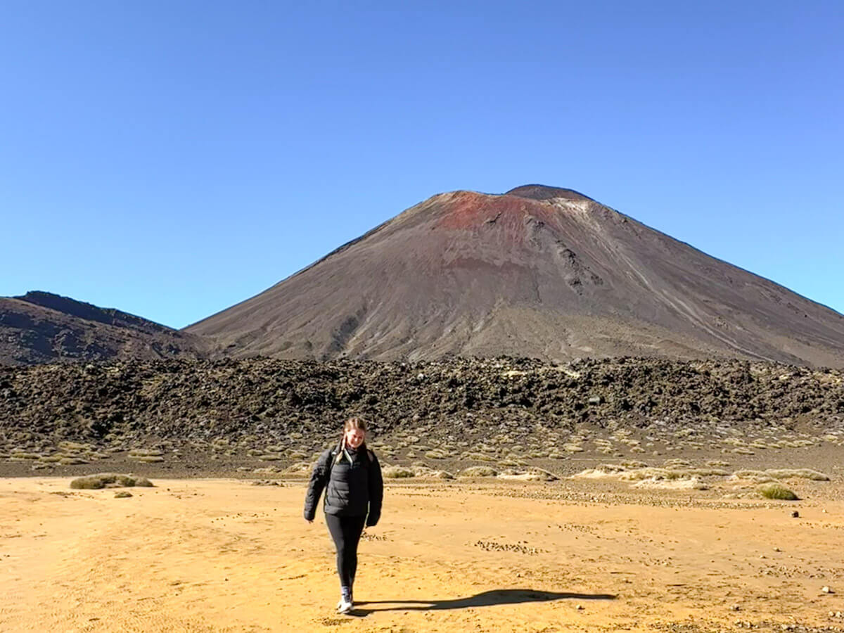 Charlie is walking across a vast, sandy volcanic plain with the prominent conical peak of Mount Ngauruhoe in the background, a key feature of the Tongariro Alpine Crossing in New Zealand.