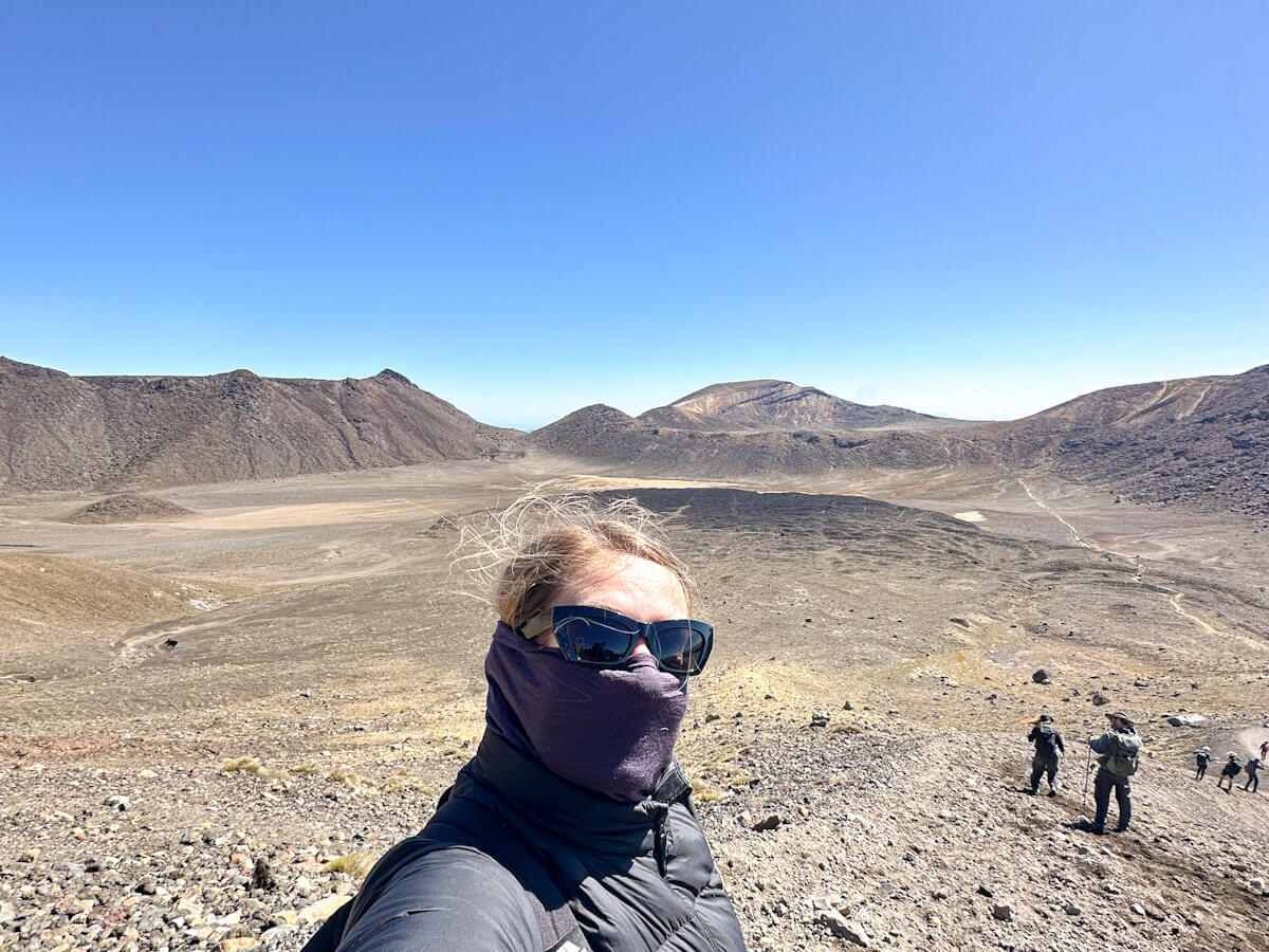 Charlie has a face covering and sunglasses on the desolate, volcanic terrain of the Tongariro Alpine Crossing, with distant mountains and craters under a clear blue sky in New Zealand